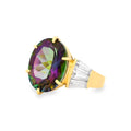 A yellow gold ring featuring a large oval faceted mystic topaz gemstone with iridescent green, purple, and pink hues, accented by baguette-cut clear crystals on the shoulders, set against a plain white background.