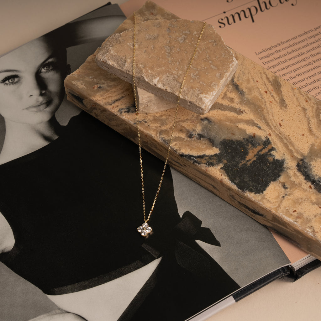 A delicate gold chain necklace with a small clear crystal pendant rests on a textured stone slab and an open magazine. 