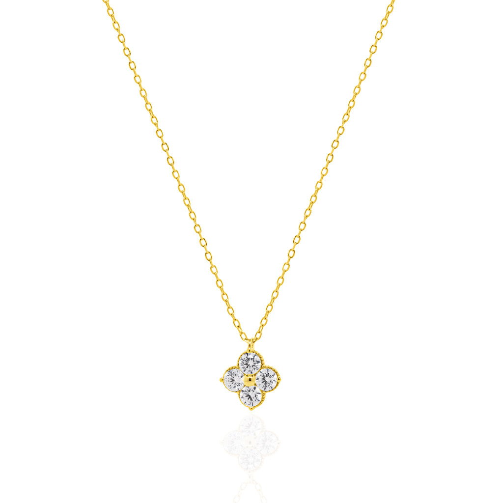 A delicate gold chain necklace with a small four-petal flower pendant set with clear gemstones, displayed on a clean white background with a soft reflection below.
