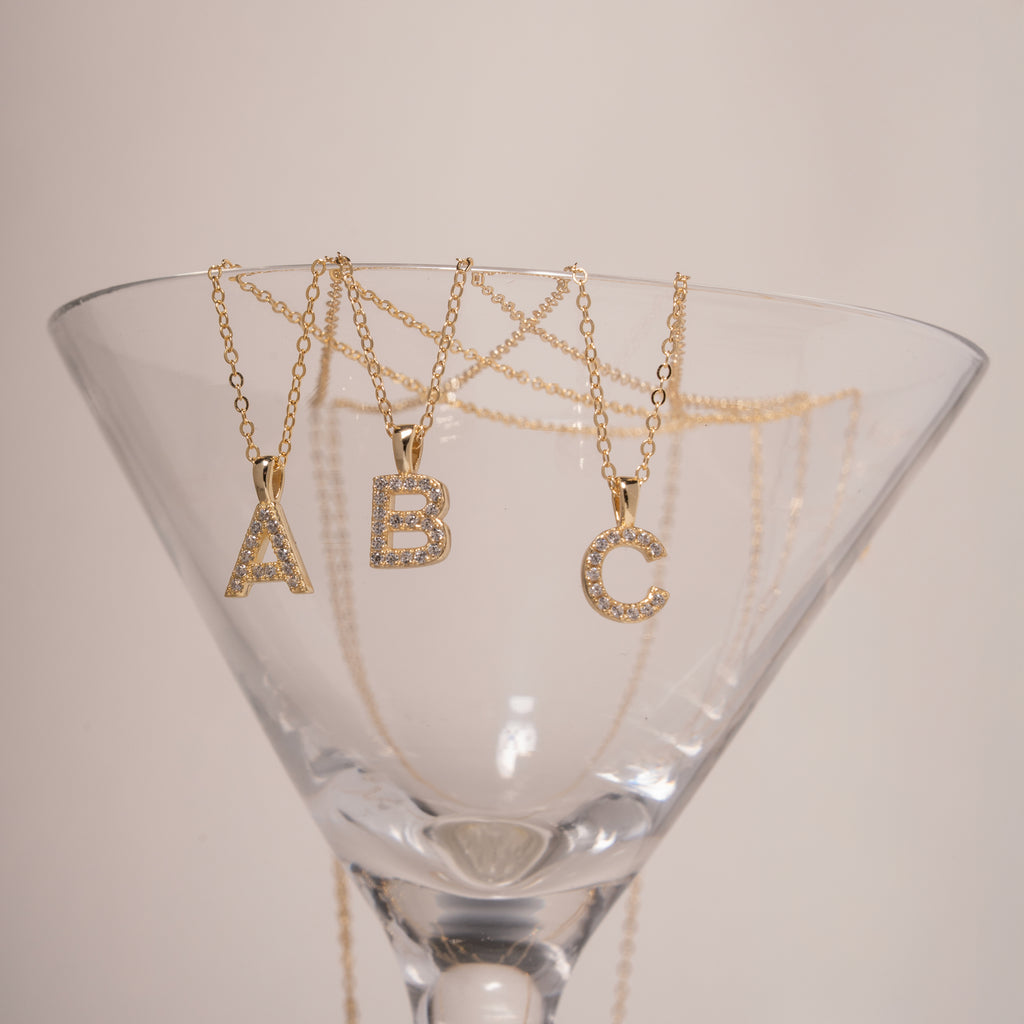 Gold tone necklaces with 'A', 'B', and 'C' clear gemstone charms in a martini glass on a neutral background