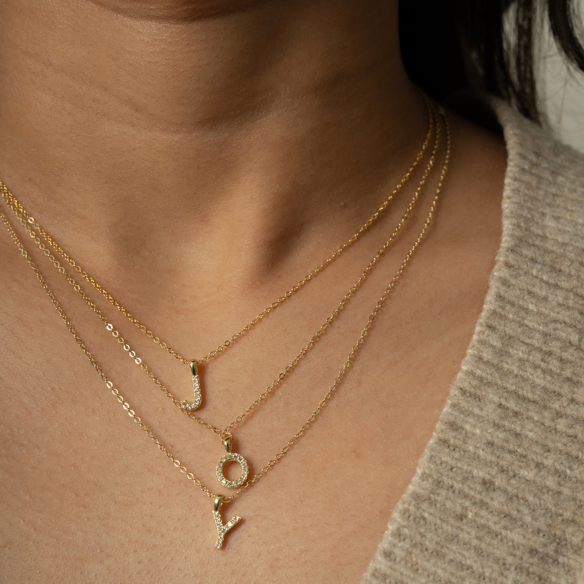 Person wearing three goldtone necklaces with clear stone initial pendants, spelling JOY