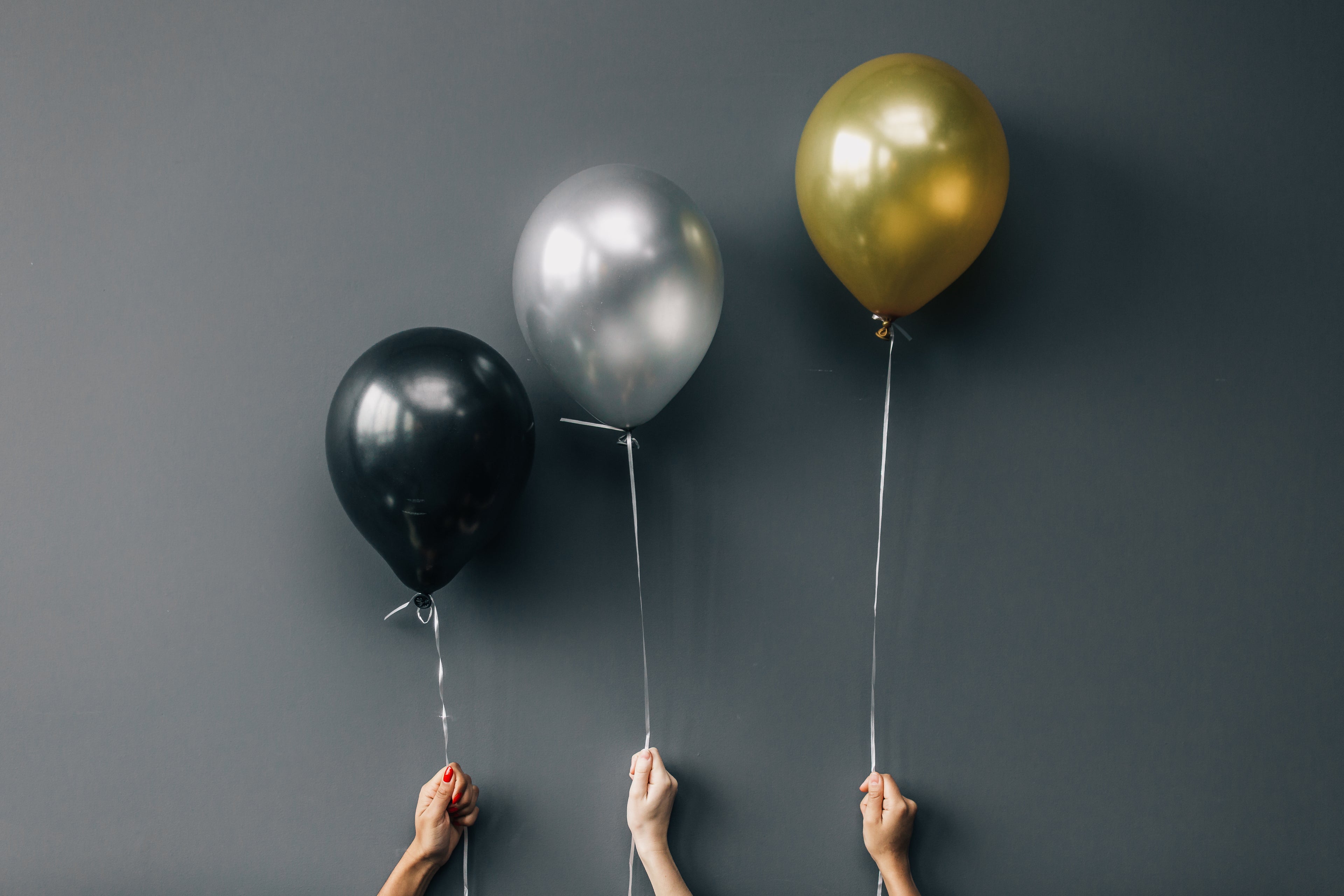 Black, gold and silver balloons floating on white ribbons against a grey background.