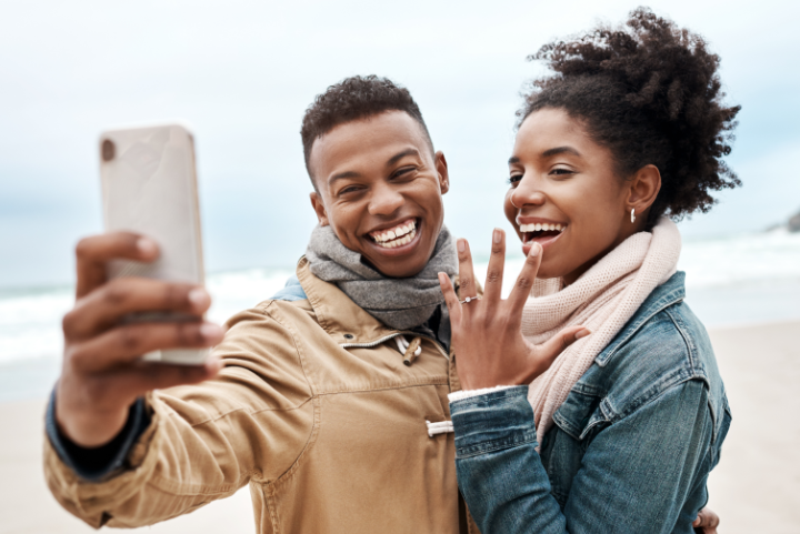 Two people taking a selfie on a beach showing off an engagement ring.