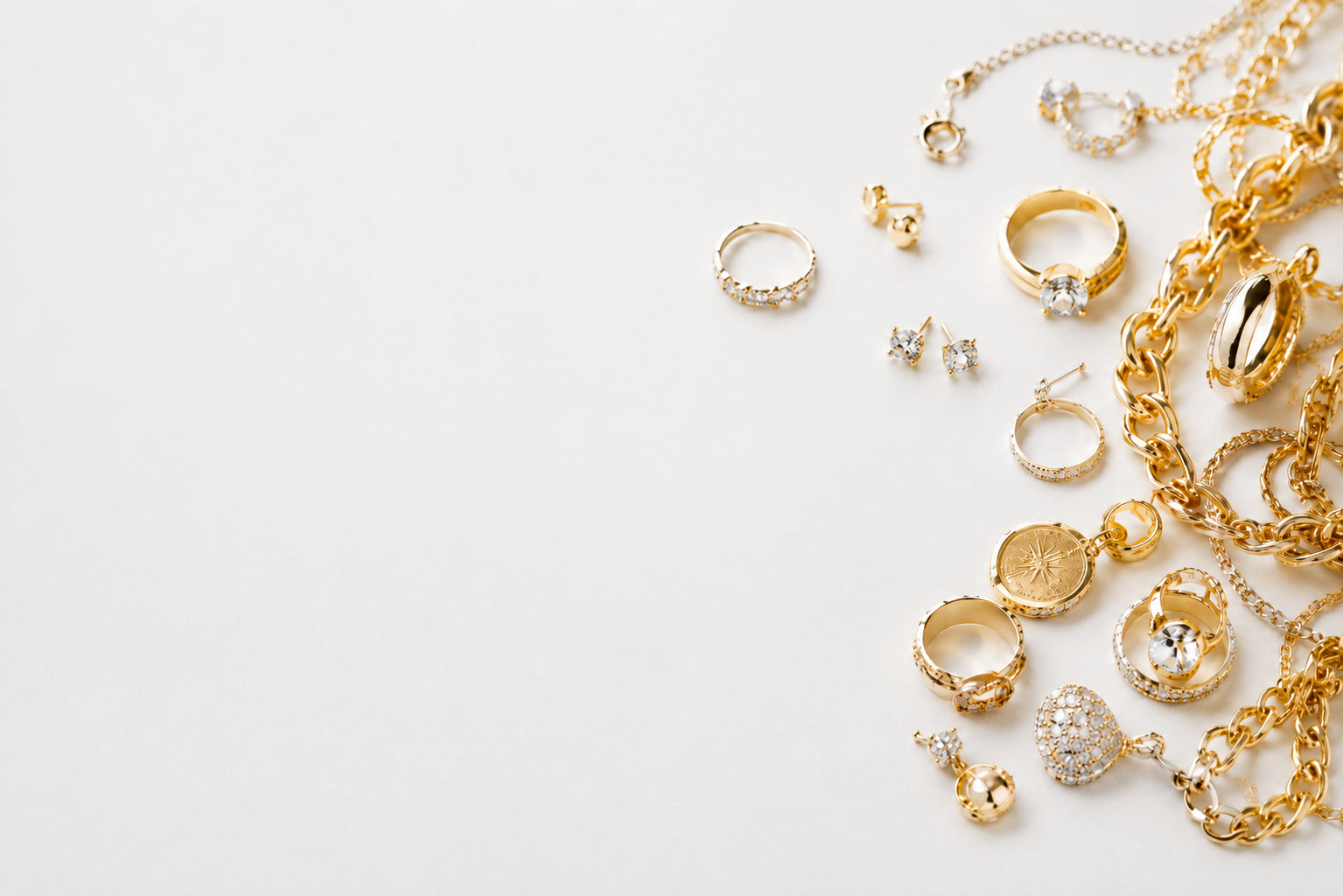 A collection of gold and silver jewelry pieces, including rings, necklaces, earrings, and bracelets, arranged on a plain white surface with negative space on the left.