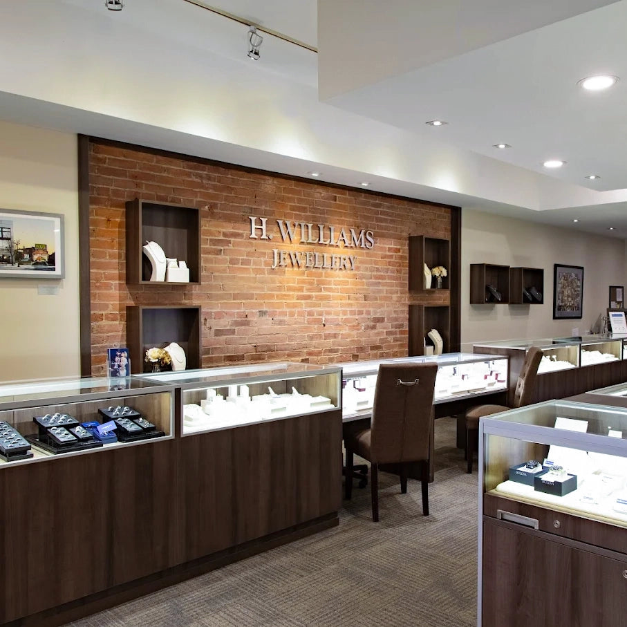 A luxury H. Williams Jewellery boutique interior featuring dark wood display cases, a brick accent wall with silver lettering, and modern lighting.