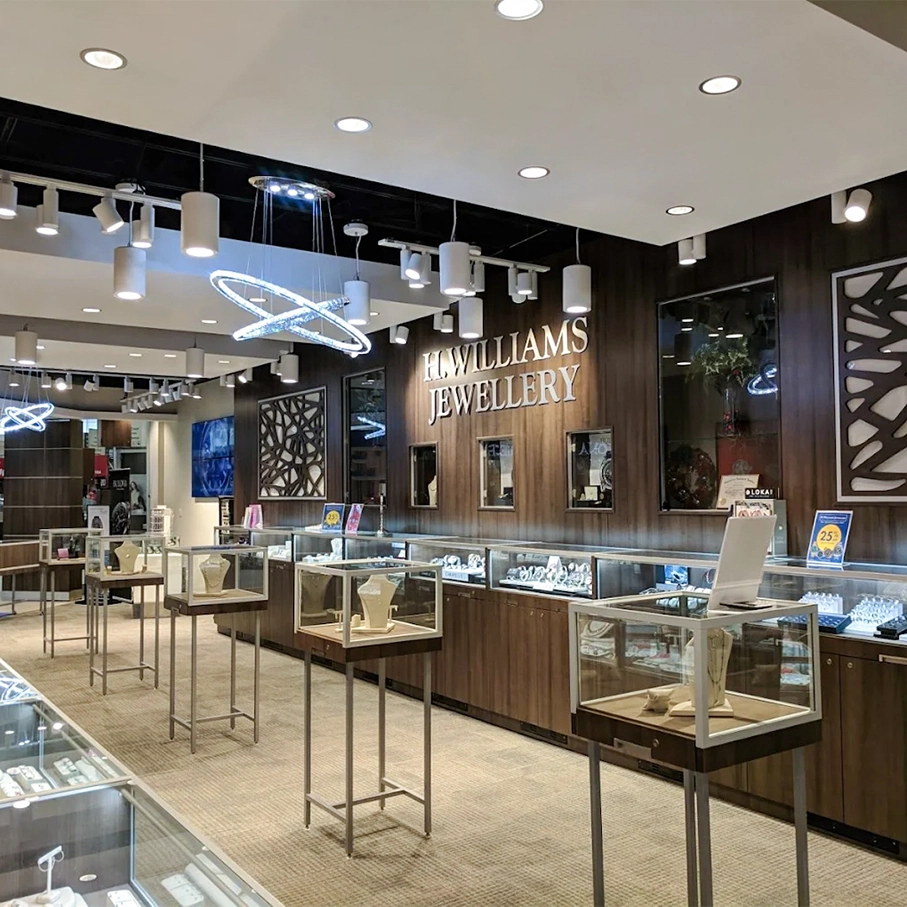 A wide interior view of a H Williams Jewellery boutique with multiple glass display cases, metal stands, pendant lights, and dark wood walls with the brand name clearly visible.