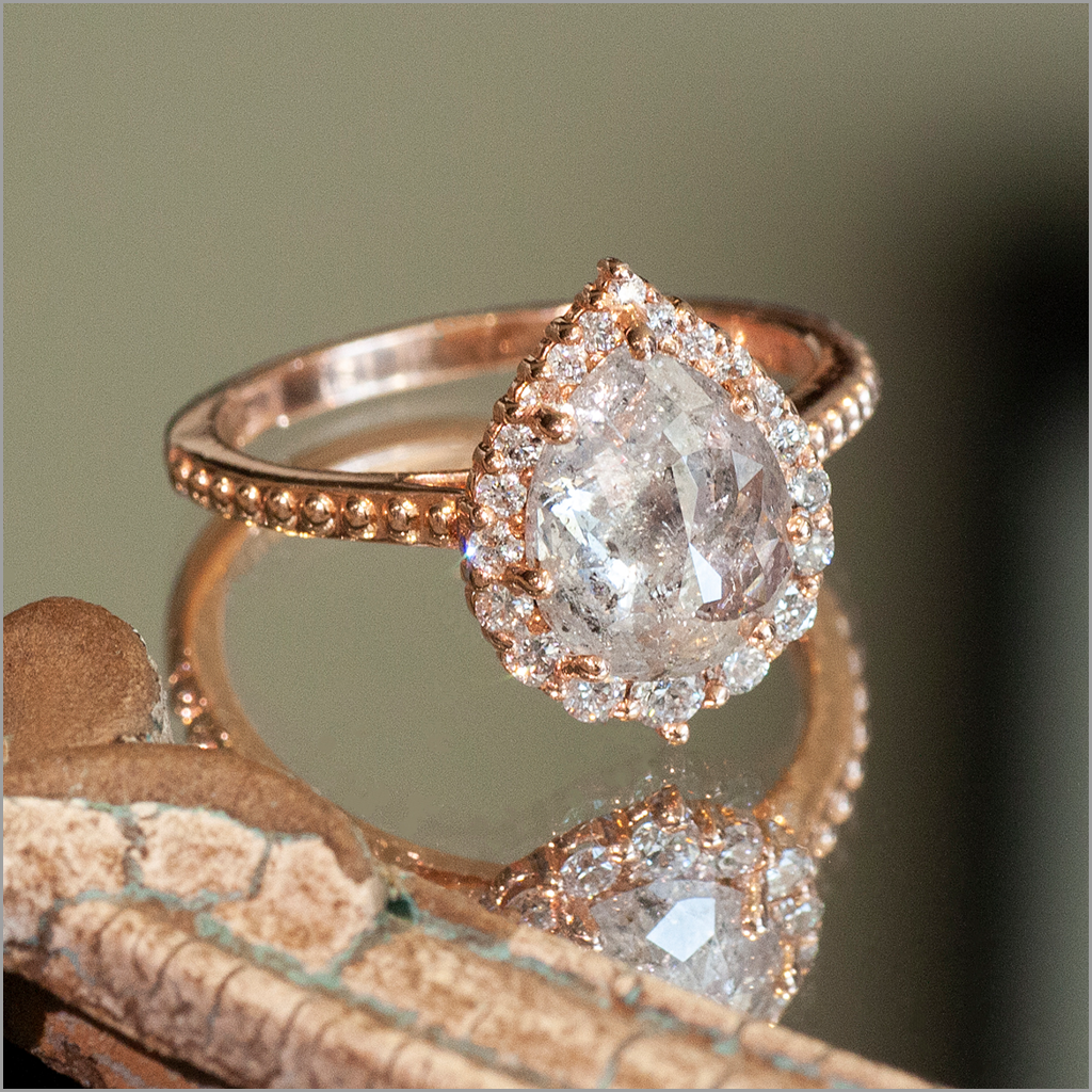 A custom-made rose gold pear-shaped rose cut diamond engagement ring with graduating halo, on a reflective surface.