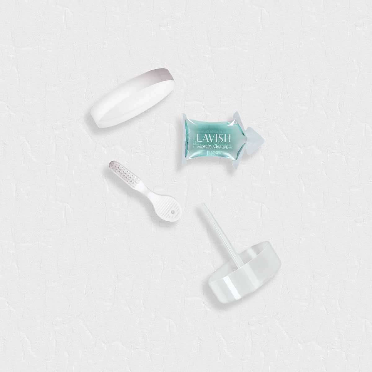 Lavish Jewellery cleaner kit components including a white cap, a small white applicator brush, a mint green sachet, and a clear soaker basket, arranged on a textured white background.