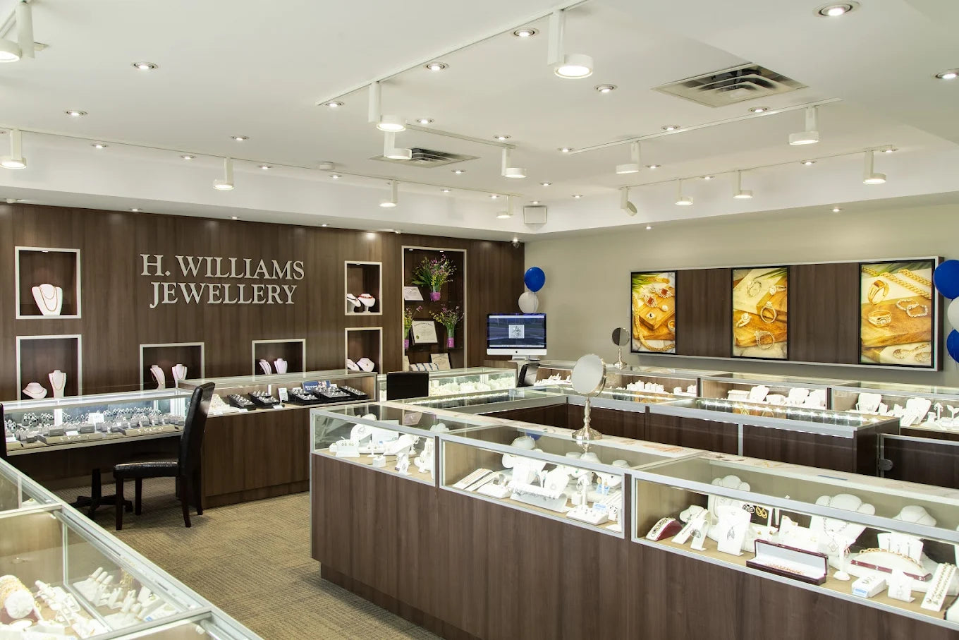 A wide interior view of an H. Williams Jewellery store with multiple glass display cases, wood-look counters, wall shelves, and jewelry items on display. Upper James Mountain location.