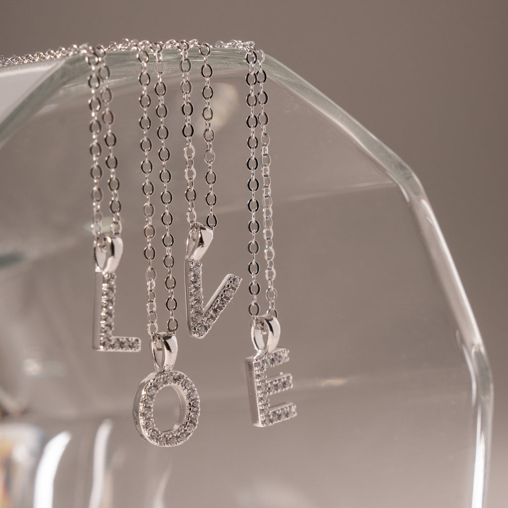 Four silver letter pendant necklaces spelling LOVE hang from a clear glass display stand against a soft gray background.