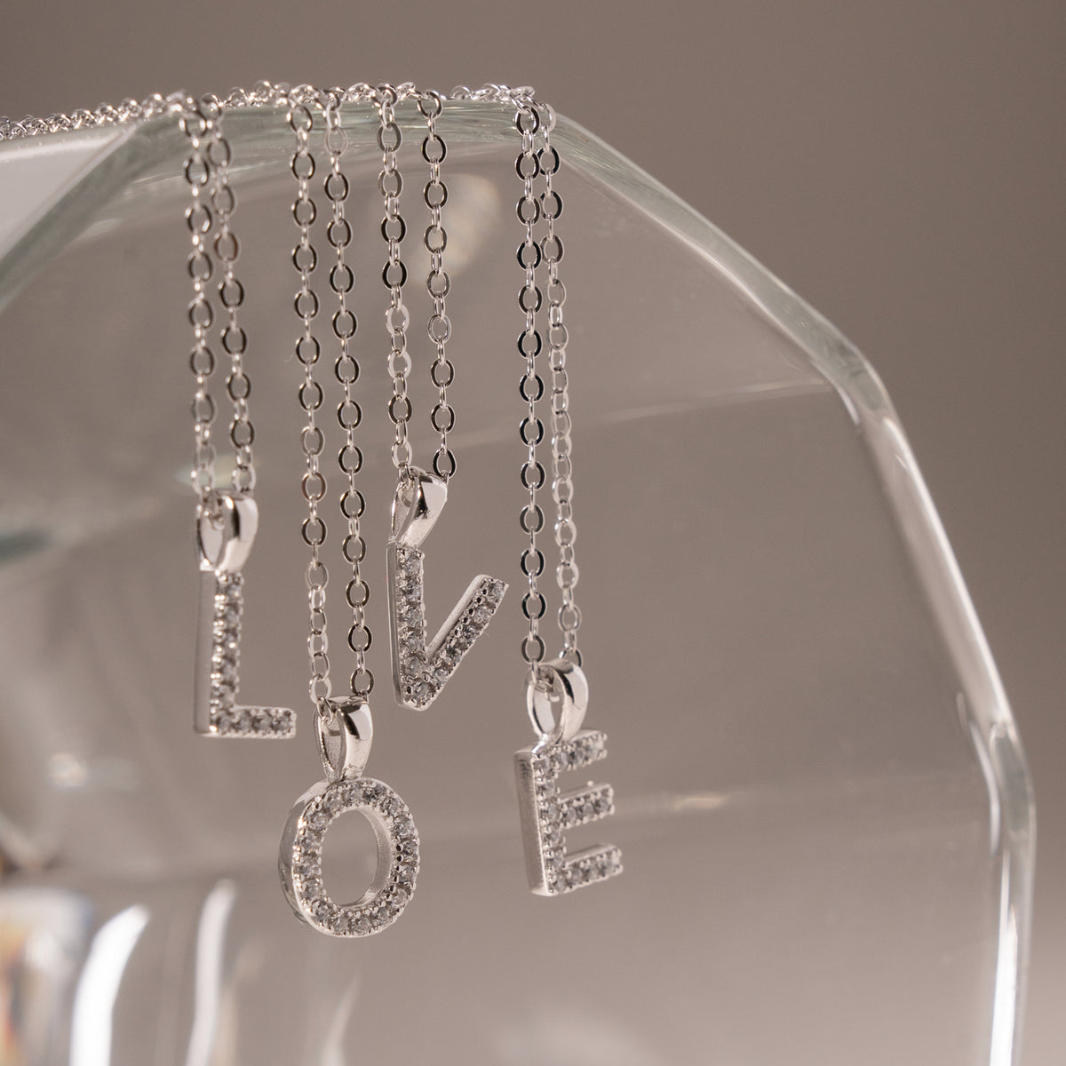 Four silver letter pendant necklaces spelling LOVE hang from a clear glass display stand against a soft gray background.