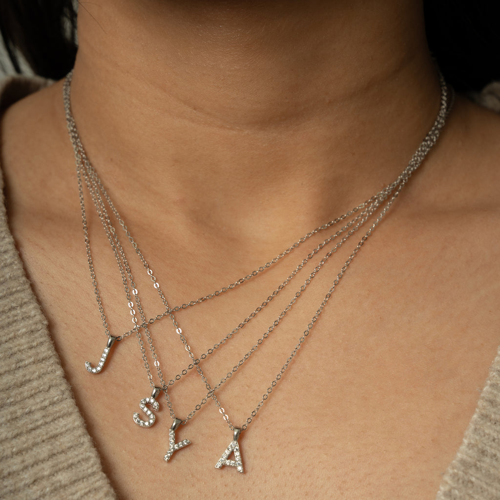 A close-up of a person's neck and upper chest wearing layered silver necklaces with letter pendants spelling JSYA, each adorned with small clear stones.