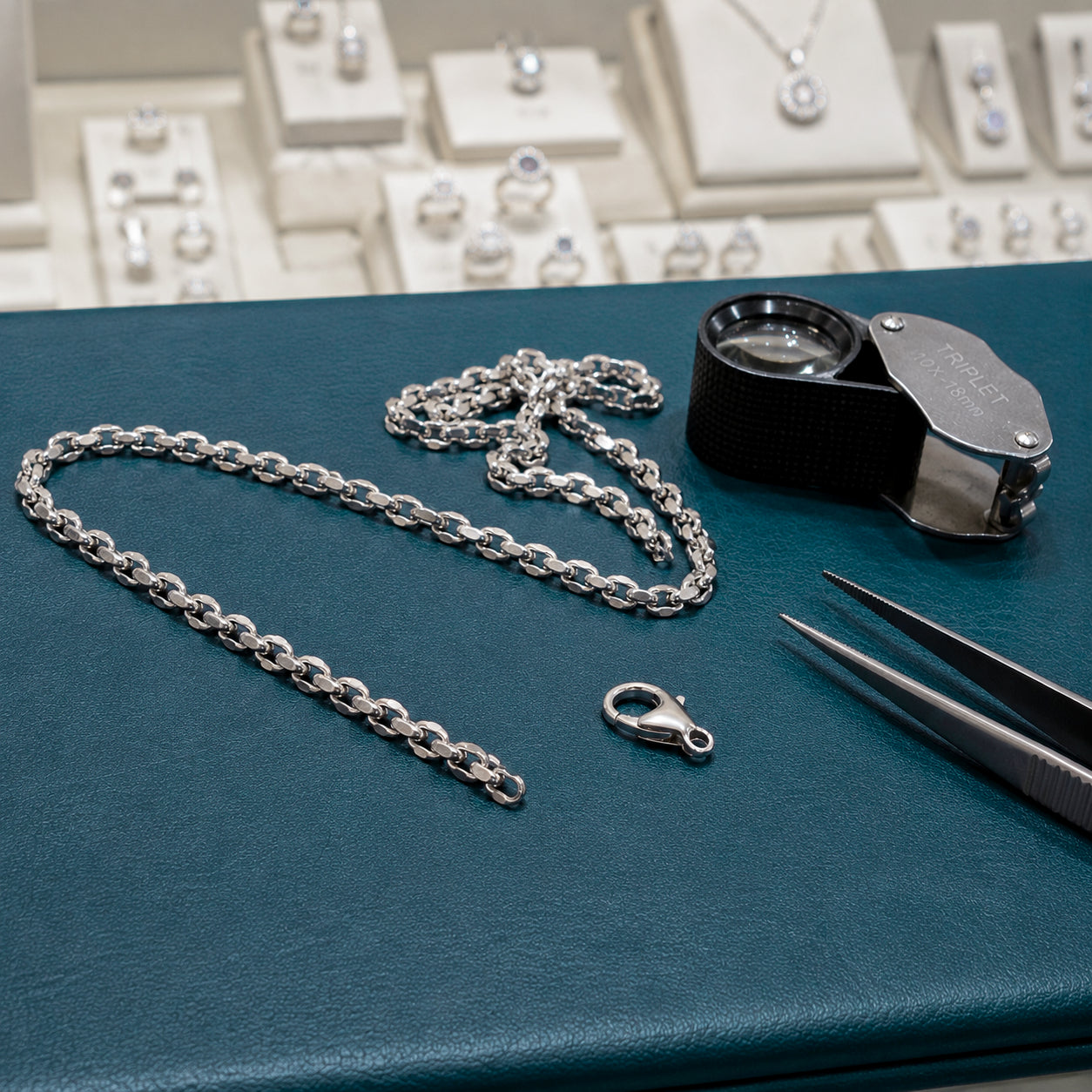 Silver chain with clasp, magnifying glass, and tweezers on a blue surface with jewelry display in the background.