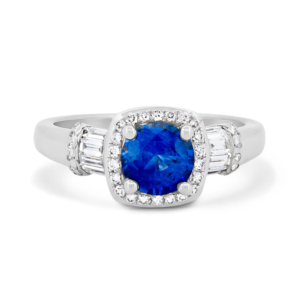 A white gold ring featuring a cushion-round blue sapphire center stone surrounded by a cushion-shaped halo of diamonds, with baguette-cut side diamonds on a white background.