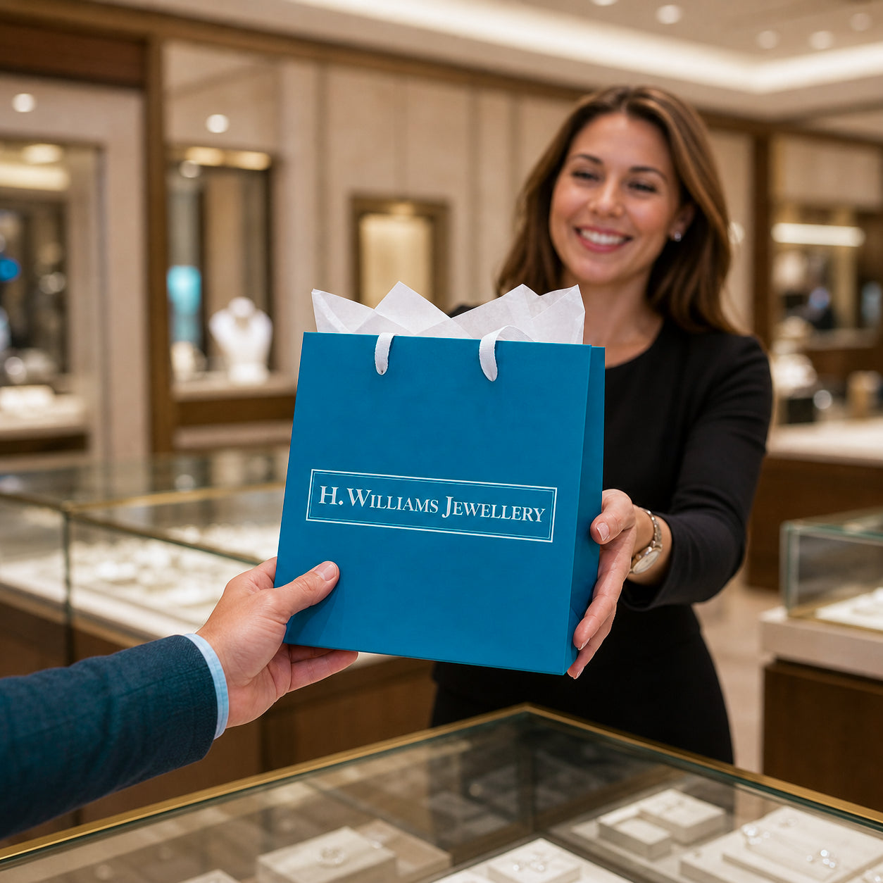 An H. Williams Jewellery shopping bag is handed by a smiling woman inside a jewelry store.