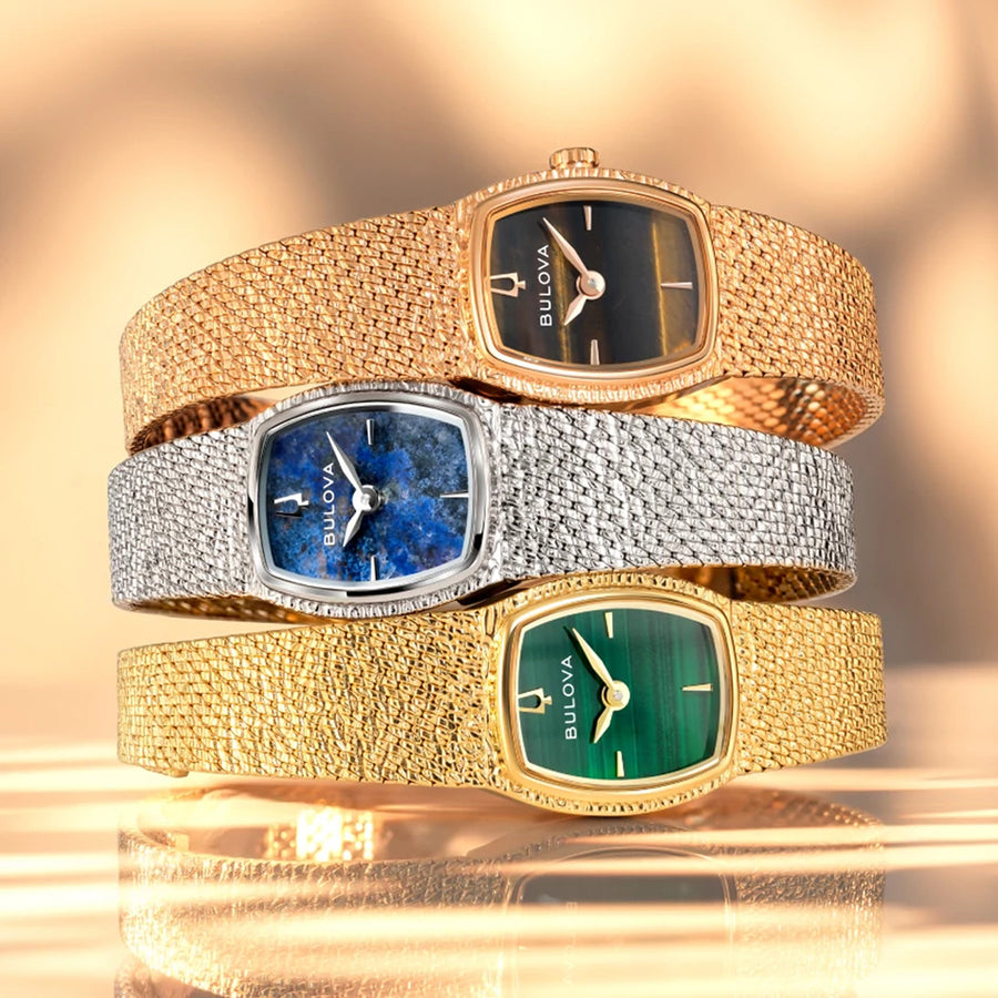 Three Bulova watches with different colored faces and gold and silver bands on a reflective surface.
